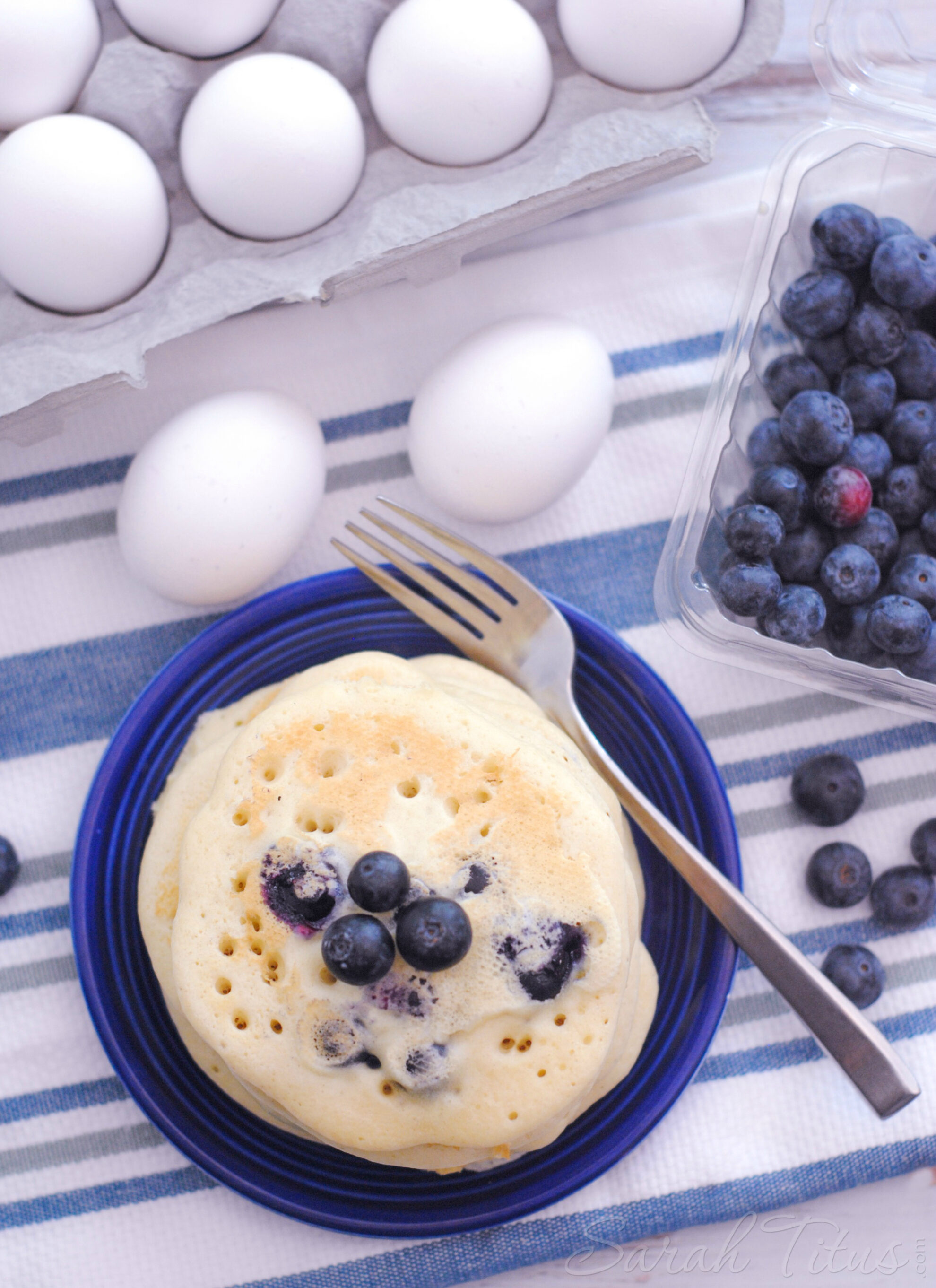 Tempting blueberry pancakes on a blue plate on a white and blue striped towel with eggs and blueberries on the side