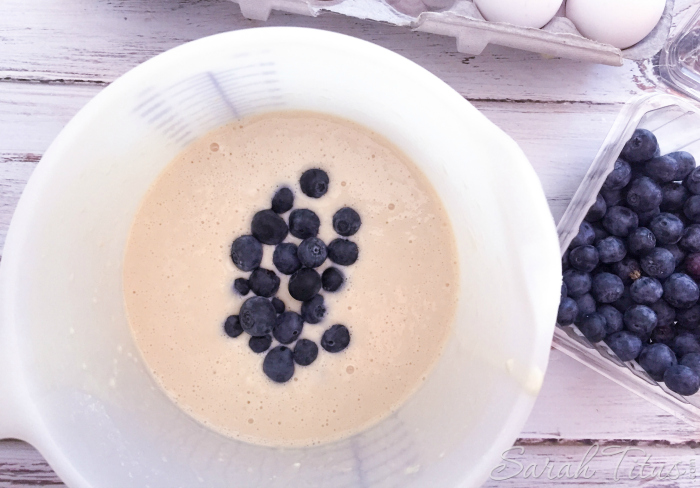 Pancake batter with fresh blueberries drizzled on the top and blueberries and eggs on the side on a white wood table