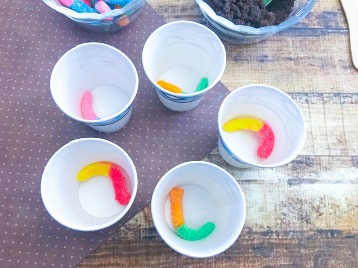 Placing gummy worms in the bottom on Dixie cups sitting on wooden table