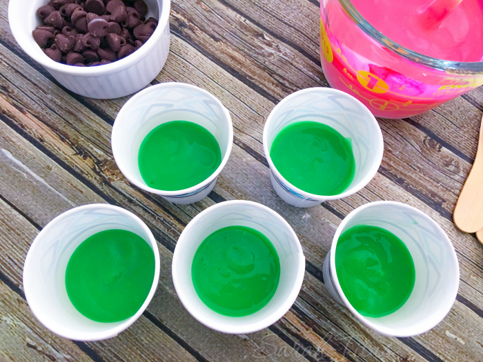 Dixie cups with green colored pudding in the bottom sitting next to chocolate chips and pink colored pudding