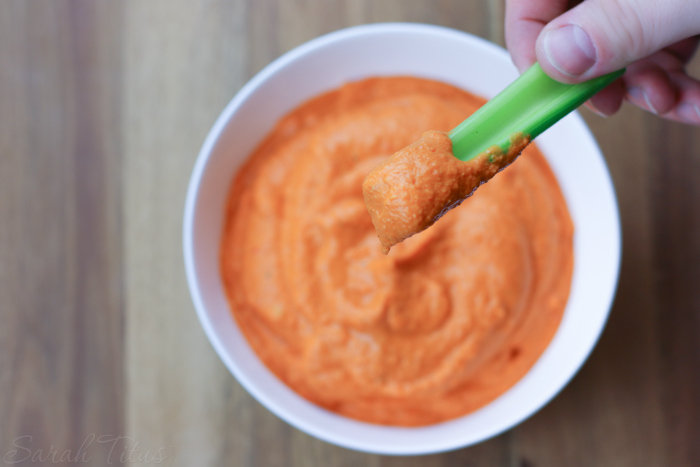 White bowl of roasted red pepper hummus sitting on a wood table and hand holding celery stick with scoop of hummus