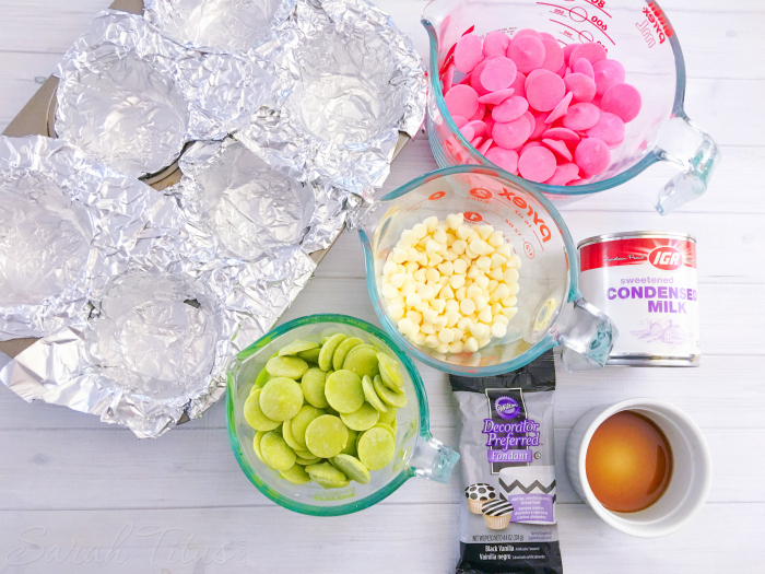 Watermelon Fudge ingredients laid out including foil lined muffin tin, green and pink melting candy, vanilla, black fondant, sweetened condensed milk and white chocolate chips