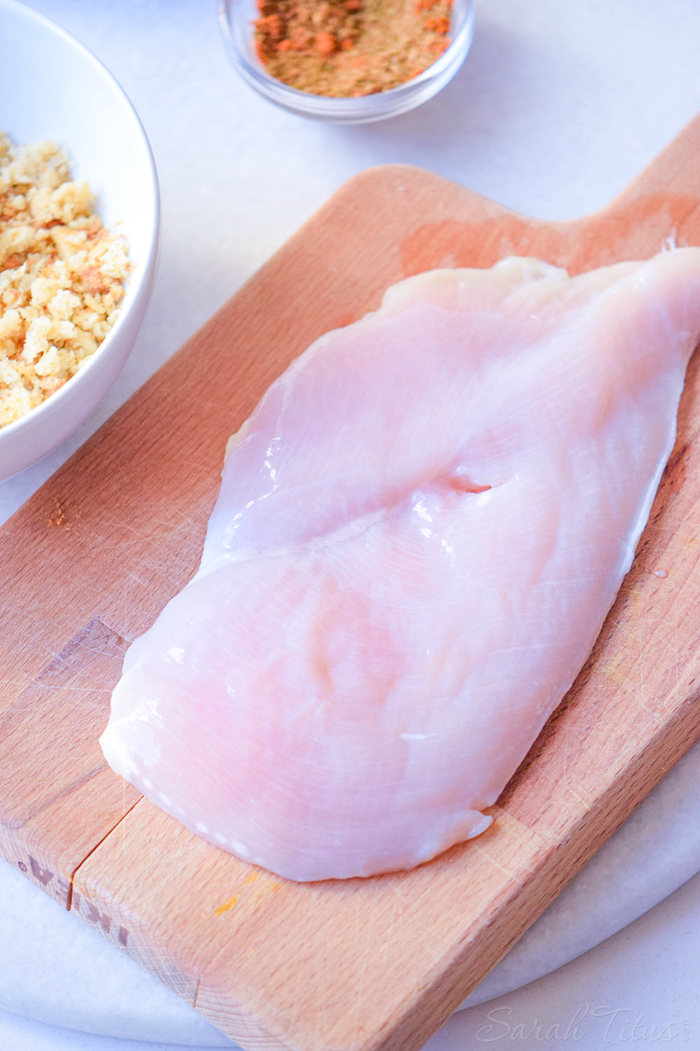 Large chicken breast on a cutting board with seasonings in bowls on the side