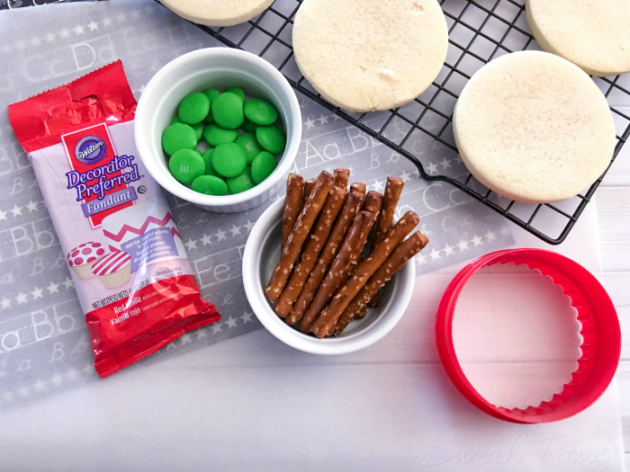 Apple Sugar Cookie ingredients: round sugar cookies on cooling tray, red fondant, green M&M's, pretzel sticks, and fluted round cookie cutter