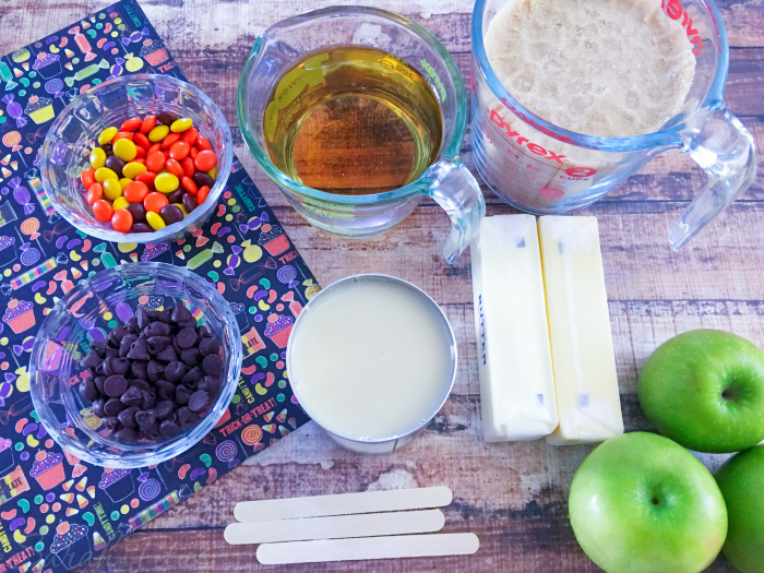 All ingredients for gourmet caramel apples laid out: apples, corn syrup, brown sugar, melted butter, sea salt, sweetened condensed milk, Reese's pieces, chocolate chips