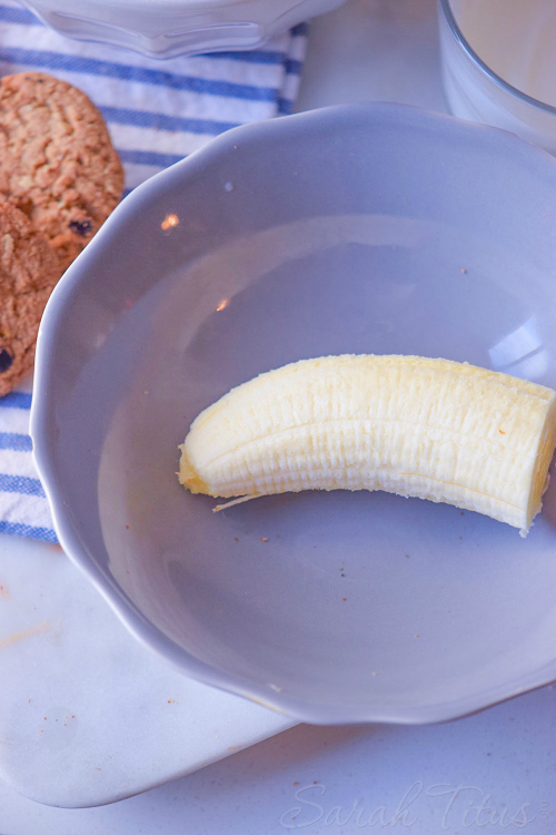 Half of a banana in a blue bowl with chocolate chip cookies and milk on the side