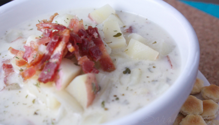 Tempting white bowl of clam chowder topped with chopped potatoes and bacon with oyster crackers on the side