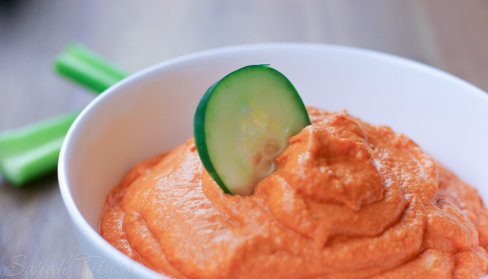 Beautiful roasted red pepper hummus in a white bowl with a slice of cucumber topping it and celery sticks on the side
