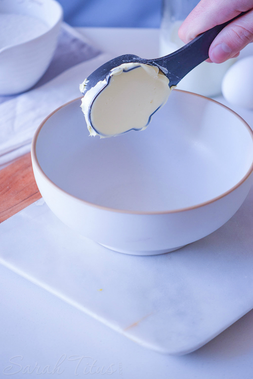 Measuring spoon full of butter being scooped into a white bowl with baking ingredients around it