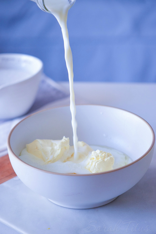 Milk pouring over butter in white bowl with baking ingredients around