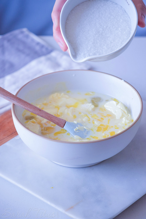 Sugar being poured over butter, milk and sugar mixture in white bowl