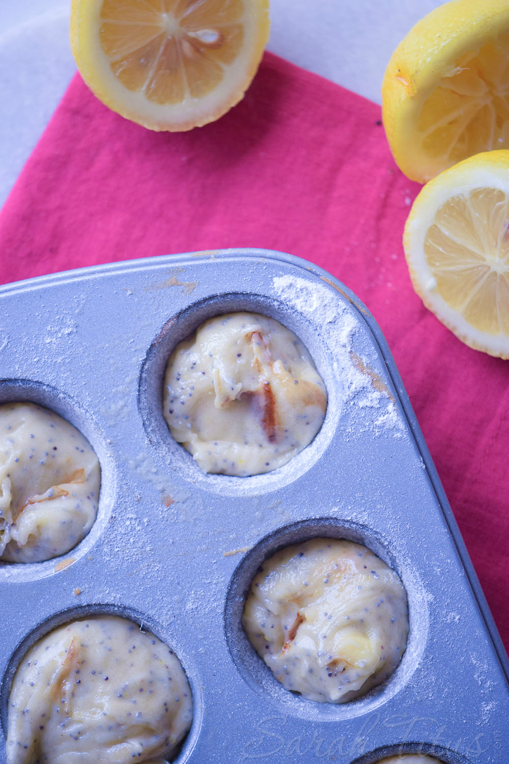 Muffin pan filled with lemon poppy batter sitting on a pink cloth napkin with fresh cut lemons on the side