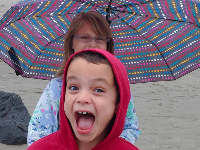 Boy photo bombing girl with colorful umbrella on a cloudy beach on a road trip