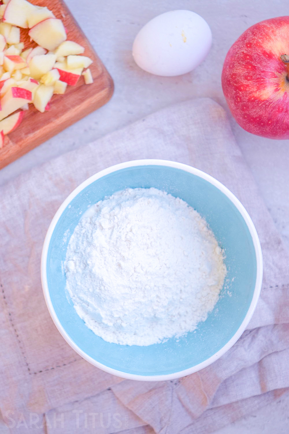 Blue bowl with dry ingredients for baked apple doughnuts with an egg, apple and chopped apples on the side