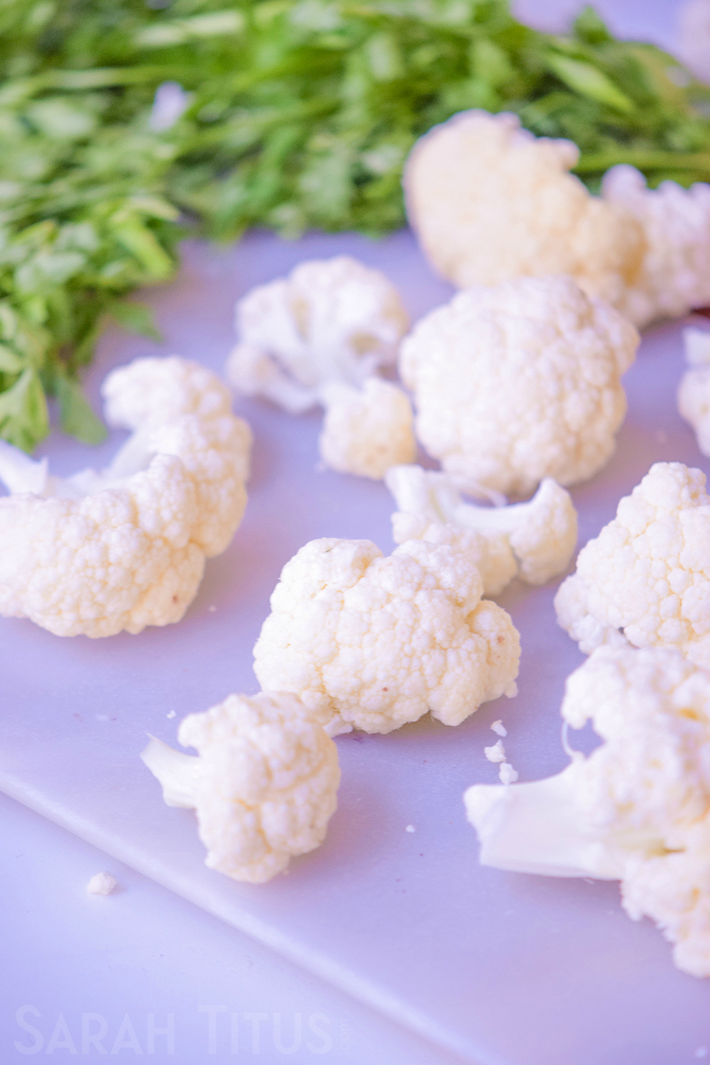 Chopped up pieces of cauliflower on a white cutting board with greenery in the background