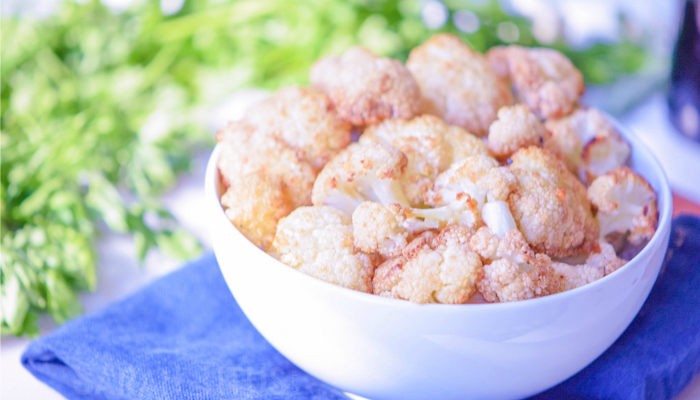  Cauliflower Bites looking very appetizing in a white bowl on a blue towel with greenery in the background