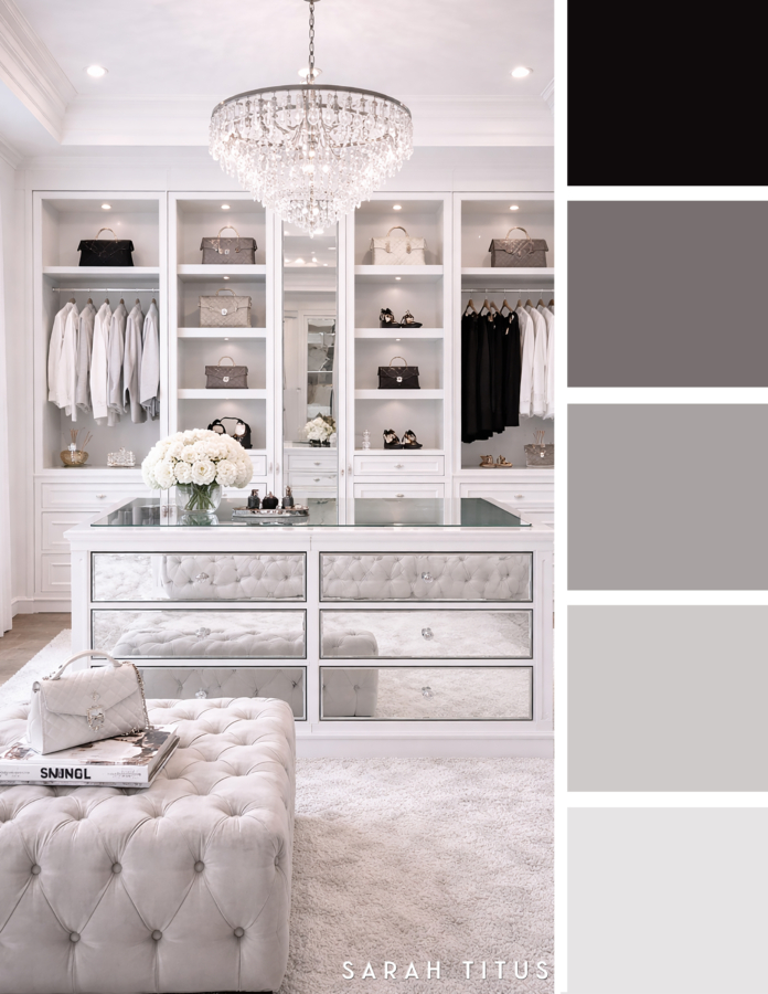 Luxe white walk in closet with mirrored drawers, crystal chandelier, tufted bench, display shelves, and soft gray handbags in a white, gray, silver, and black color palette.