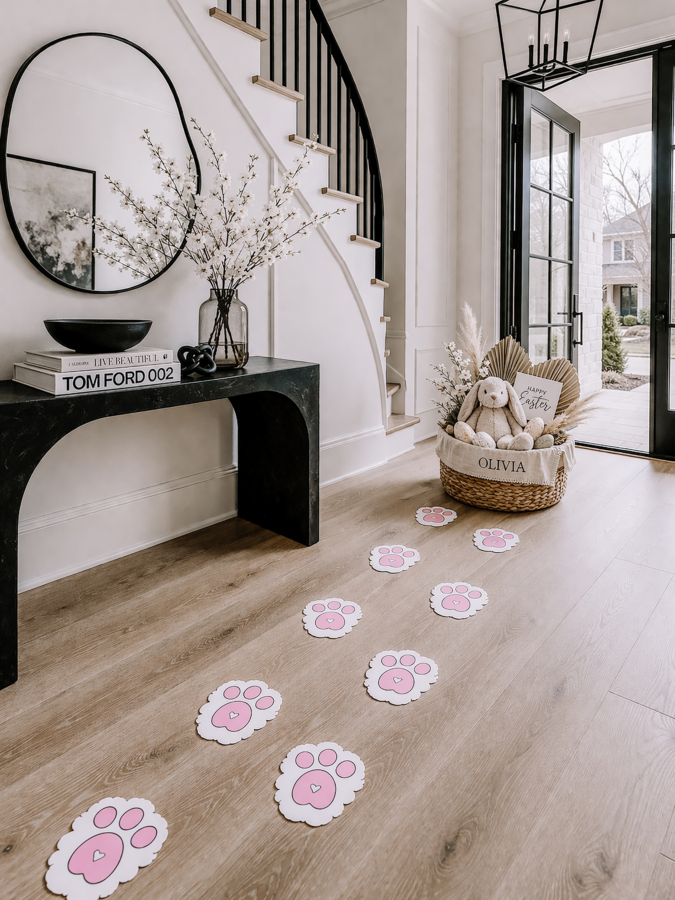 Pink printable Easter bunny paw prints scattered across a light wood floor in a modern black and white entryway, leading to a personalized Easter basket filled with a stuffed bunny and Easter decor.