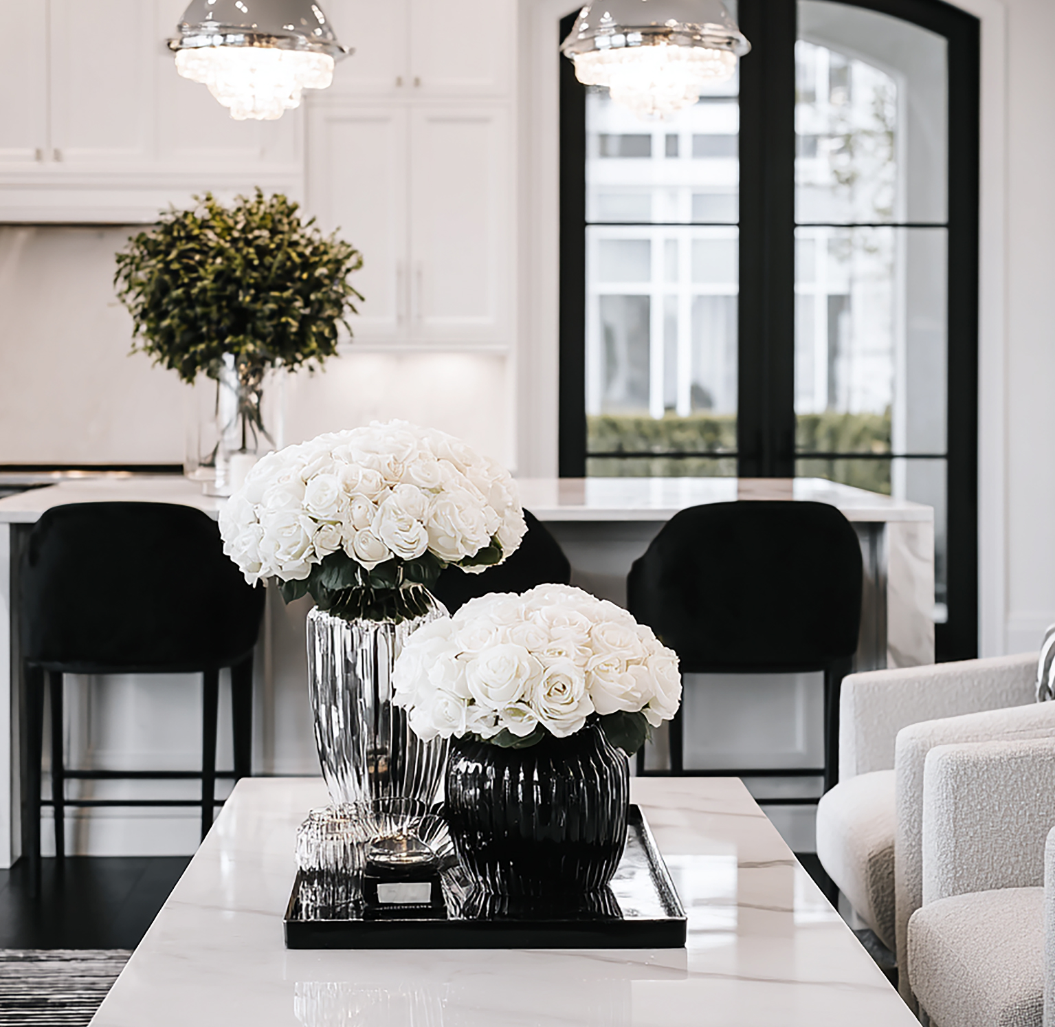 Bright modern black and white living room with white roses, crystal vases, a sleek coffee table, black barstools, and arched black framed windows.