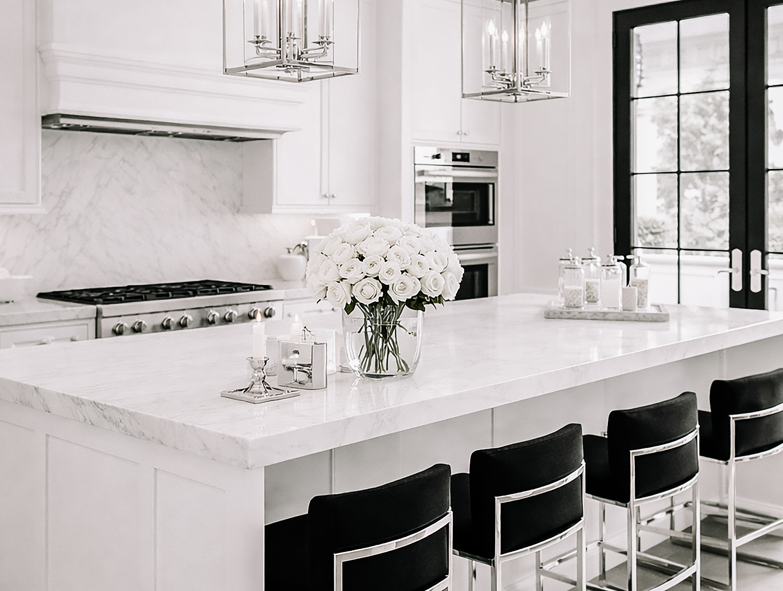 Black, white, and silver luxury kitchen with a marble island, black barstools, white roses, pendant lanterns, and black French doors