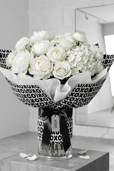 White flower bouquet wrapped in black and white XOXO Mother’s Day flower wrapping paper with white tissue paper, a black ribbon, and a matching tag in a glass vase.