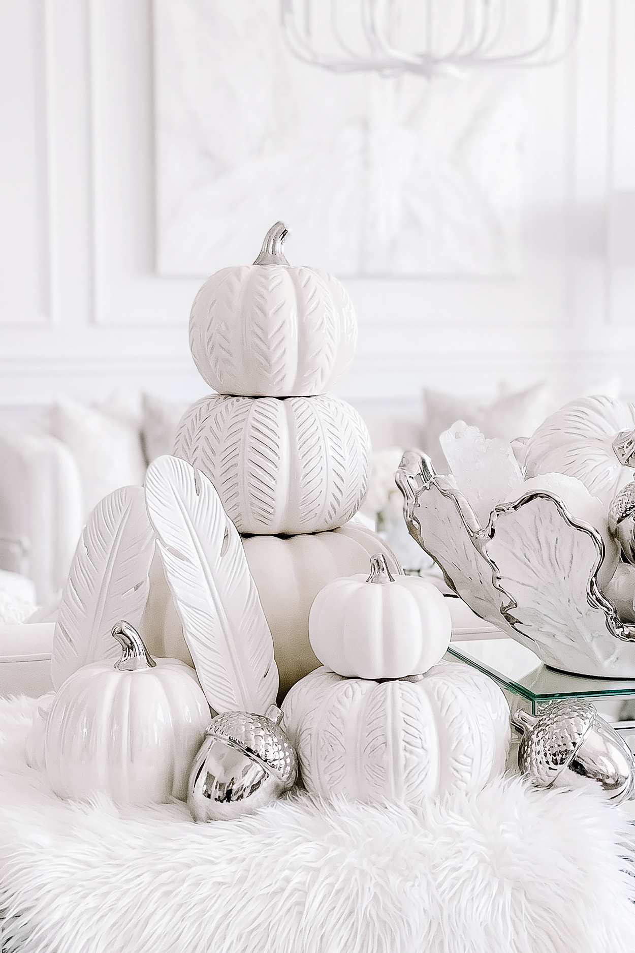 White and silver fall decor display with stacked white pumpkins, feather accents, silver acorns, and a decorative bowl on a fluffy white table runner