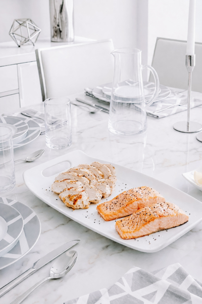 Grilled salmon and sliced chicken on a white serving platter set on a bright marble dining table with silver accents.