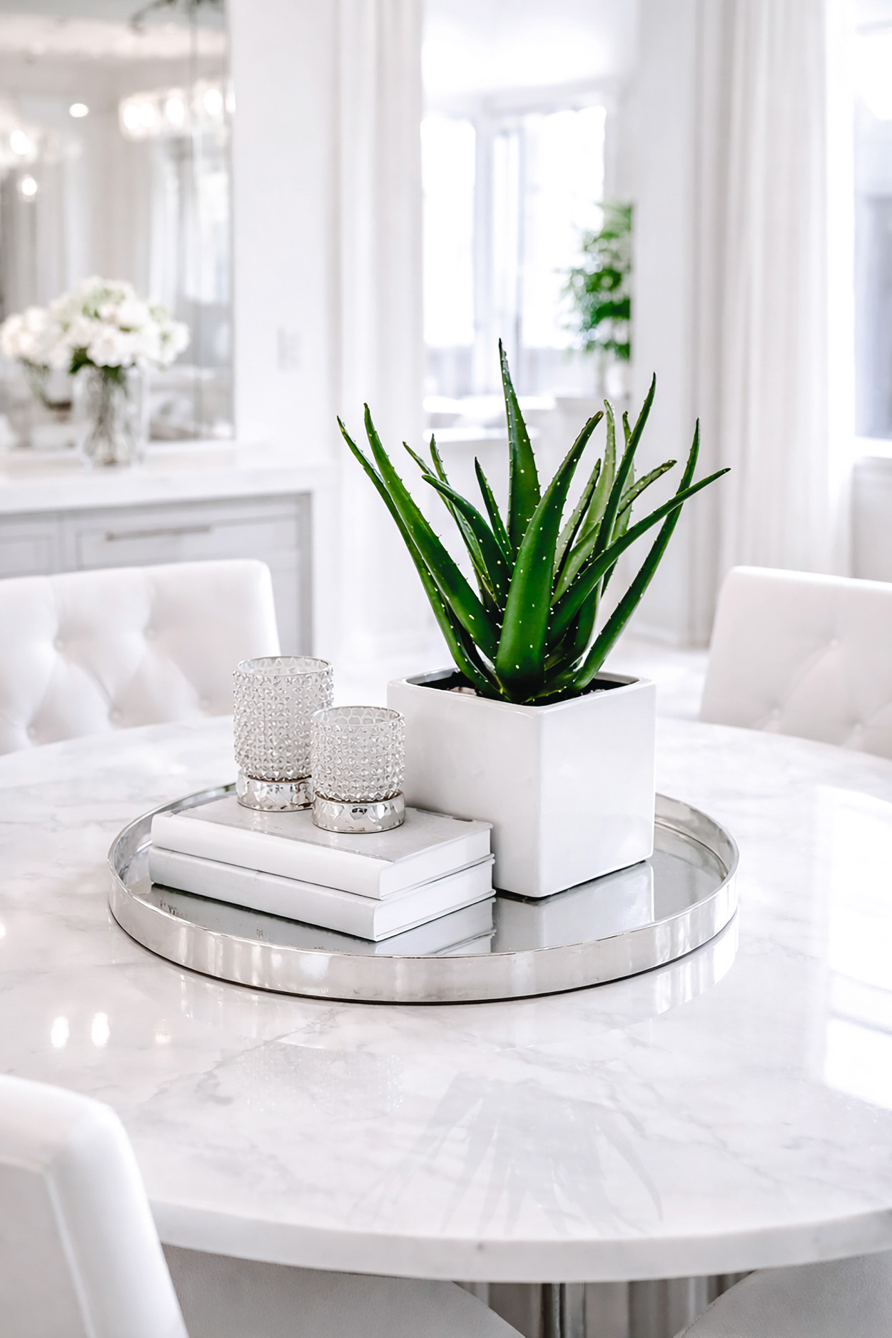 Aloe plant in a square white planter styled on a round marble dining table in a bright Z Gallerie inspired dining room with silver tray accents and chic white tufted chairs.