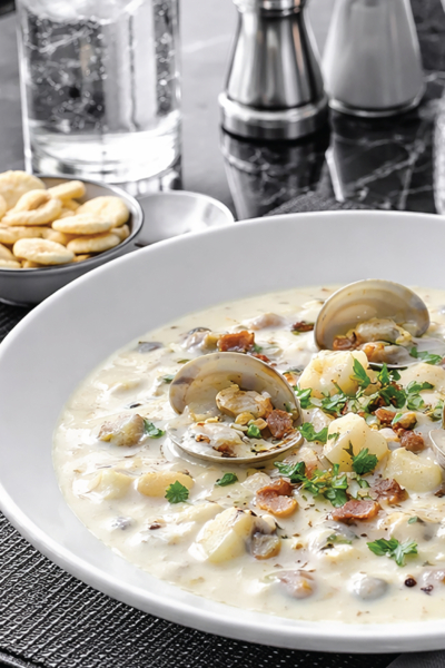 Creamy clam chowder with clams, potatoes, and herbs in a white bowl on a black marble table with oyster crackers nearby.