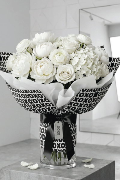 White flower bouquet wrapped in black and white XOXO Mother’s Day flower wrapping paper with white tissue paper, a black ribbon, and a matching tag in a glass vase.
