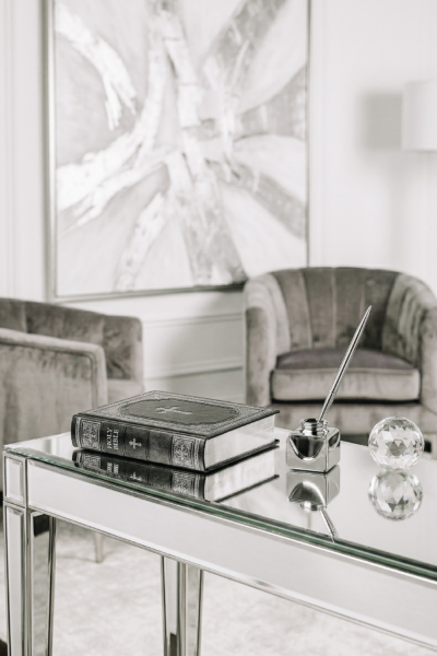 A closed Holy Bible resting on a mirrored table with a silver inkwell pen and crystal decor in a modern gray and white living room for exploring Bible study topics.
