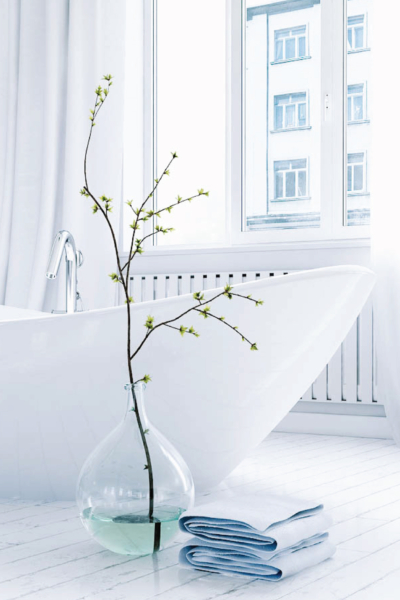 A pristine, modern white bathroom featuring a sleek freestanding bathtub with a polished silver faucet, a large clear glass vase holding a budding green branch, and neatly folded light blue towels resting on a white marble floor.