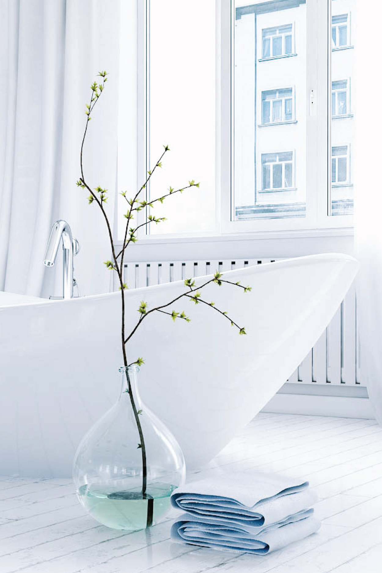 A pristine, modern white bathroom featuring a sleek freestanding bathtub with a polished silver faucet, a large clear glass vase holding a budding green branch, and neatly folded light blue towels resting on a white marble floor.